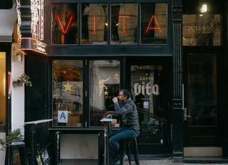 Outside of Caffe Vita's Lower East Side cafe featuring Vita signage in the windows and a customer drinking coffee while sitting on a stool.