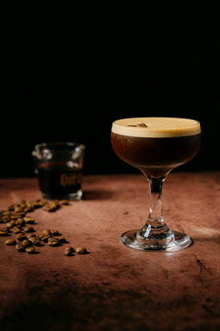 Espresso martini in coup on counter with coffee beans and shot glass of espresso